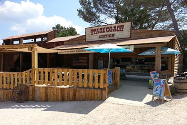 OK CORRAL propose une ambiance Far West authentique avec sa station de restauration, parfaite pour une pause en famille. Ce parc d’attractions dans le Sud de la France offre un cadre convivial où les familles peuvent se détendre tout en savourant des en-cas. Les décorations en bois et les parasols créent une atmosphère immersive, idéale après une journée d'attractions pour enfants et de sensations fortes. Profitez d'un moment de divertissement en Provence, entouré de paysages naturels
